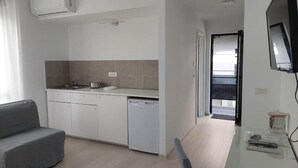 Una cucina moderna con mobili bianchi, lavastoviglie e forno integrato. Nell'area living, un divano grigio e una TV a schermo piatto montata a parete.