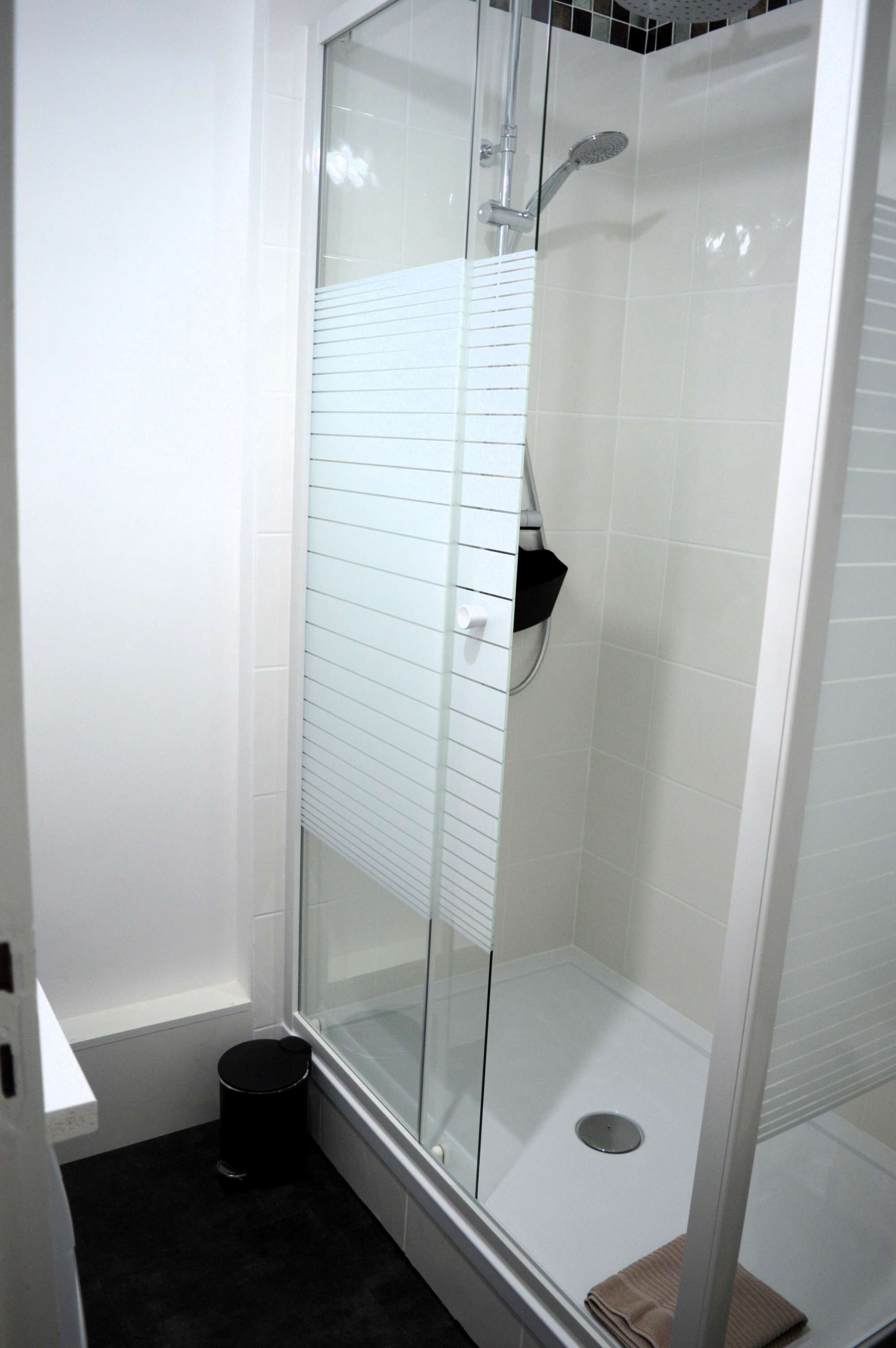 A glass-enclosed shower with white tiled walls and a black trash can.