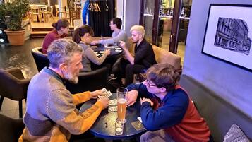 Een groep mensen speelt kaarten aan een ronde tafel in een gezellige ruimte, met potplanten en een lijst op de muur.