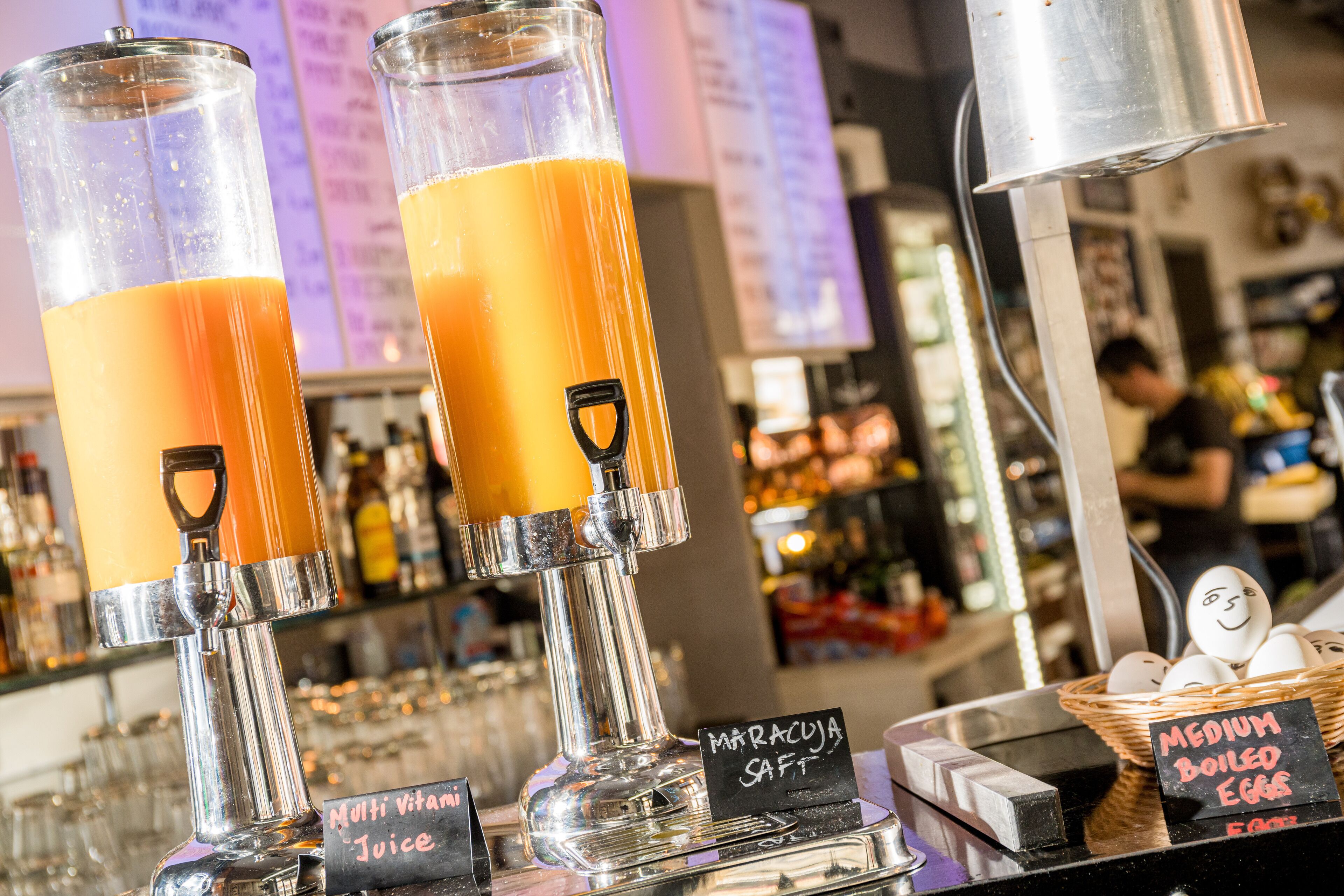 Angolo bar con due bicchieri di succo d'arancia, un cestino di uova medie e un cartello che indica 'Succo Multivitaminico'.