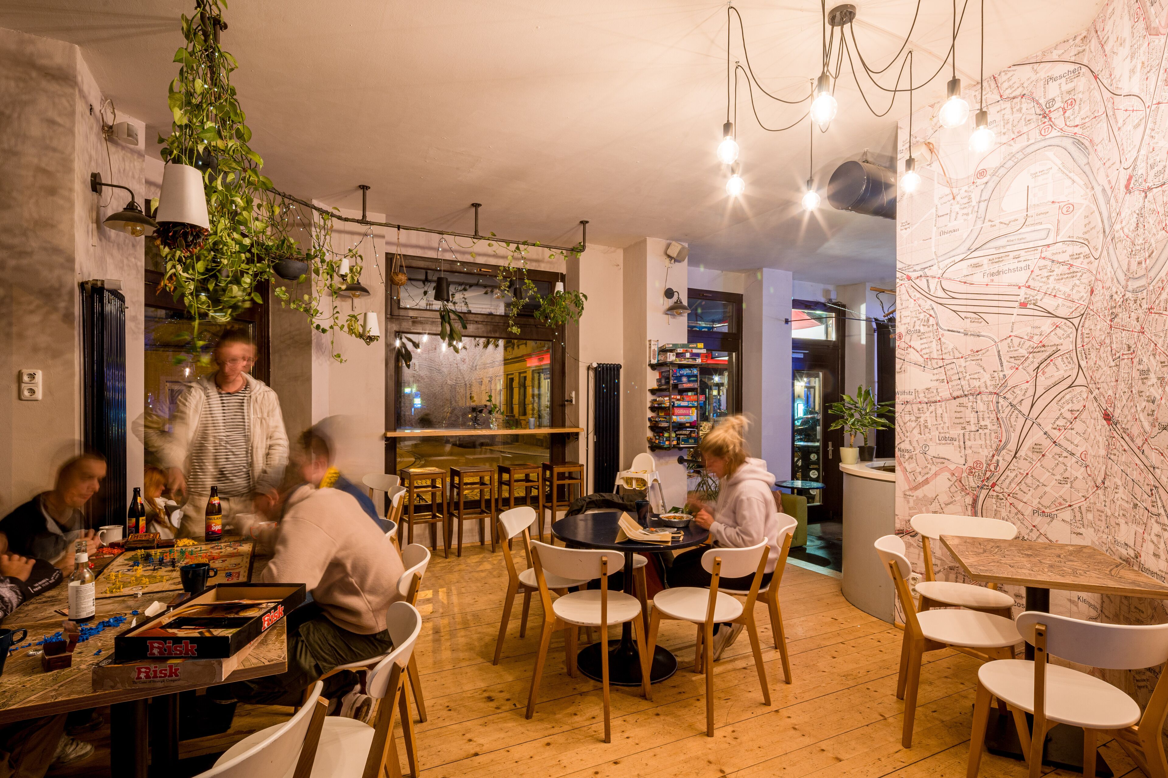 En hyggelig restaurant med træborde og stole, hængende planter og et kort på væggen.
