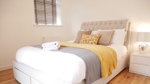 Een net opgemaakt bed met een grijs hoofdbord, wit beddengoed en een gele plaid.