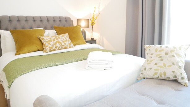 Een net opgemaakt bed met een groene plaid, twee mosterdgele kussens en een wit kussen met een bladmotief.