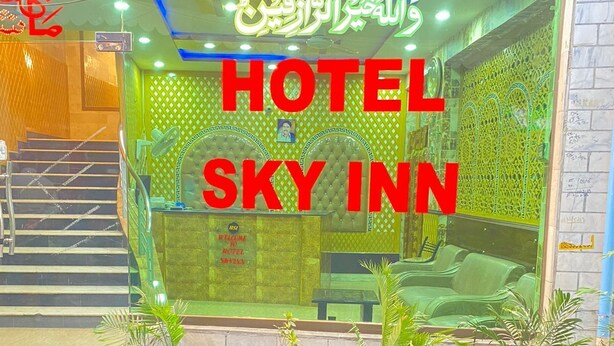 A hotel entrance with a sign reading "HOTEL SKY INN" and Arabic script above. There are steps leading up to the entrance and potted plants in front.