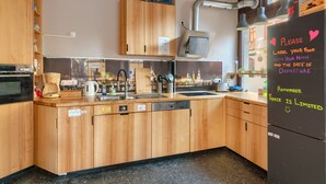 Une cuisine moderne avec des meubles en bois, un four encastré, une machine à café et un évier. Un mot est affiché sur le réfrigérateur, demandant aux invités d’indiquer leur nom et la date de leur départ sur les aliments qu’ils y placent.