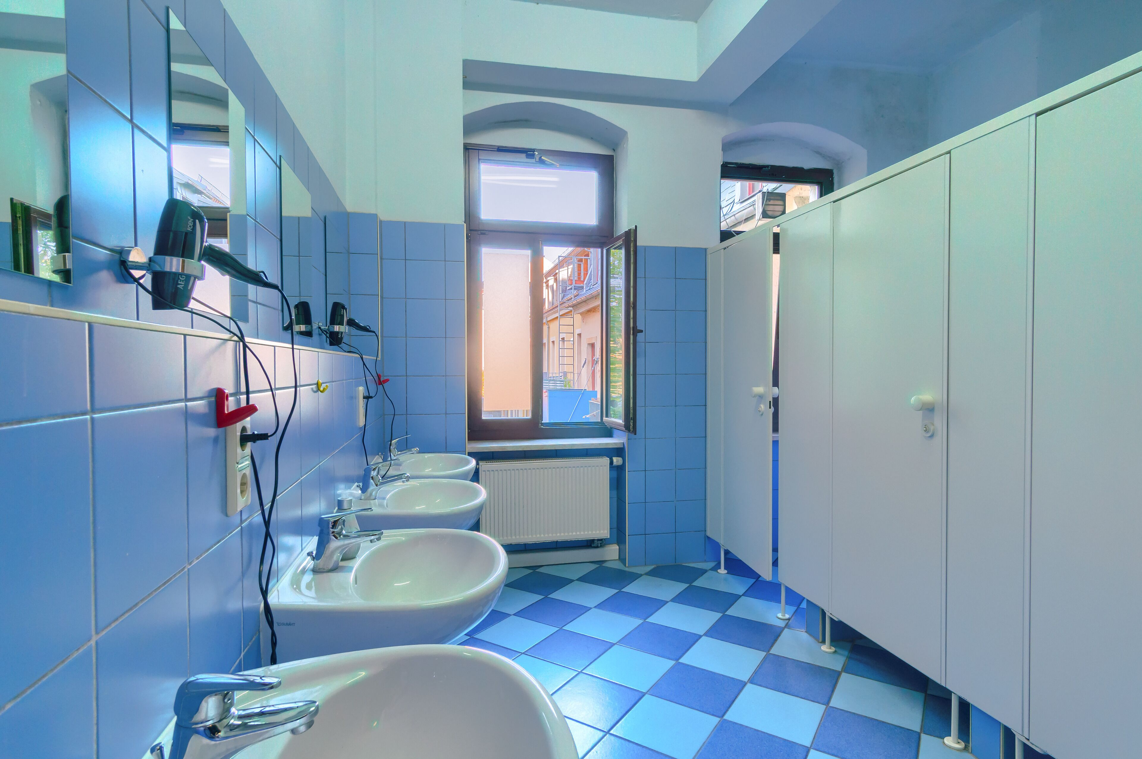 Une salle de bain avec un sol à carreaux bleus, des lavabos blancs et une fenêtre donnant sur des immeubles.