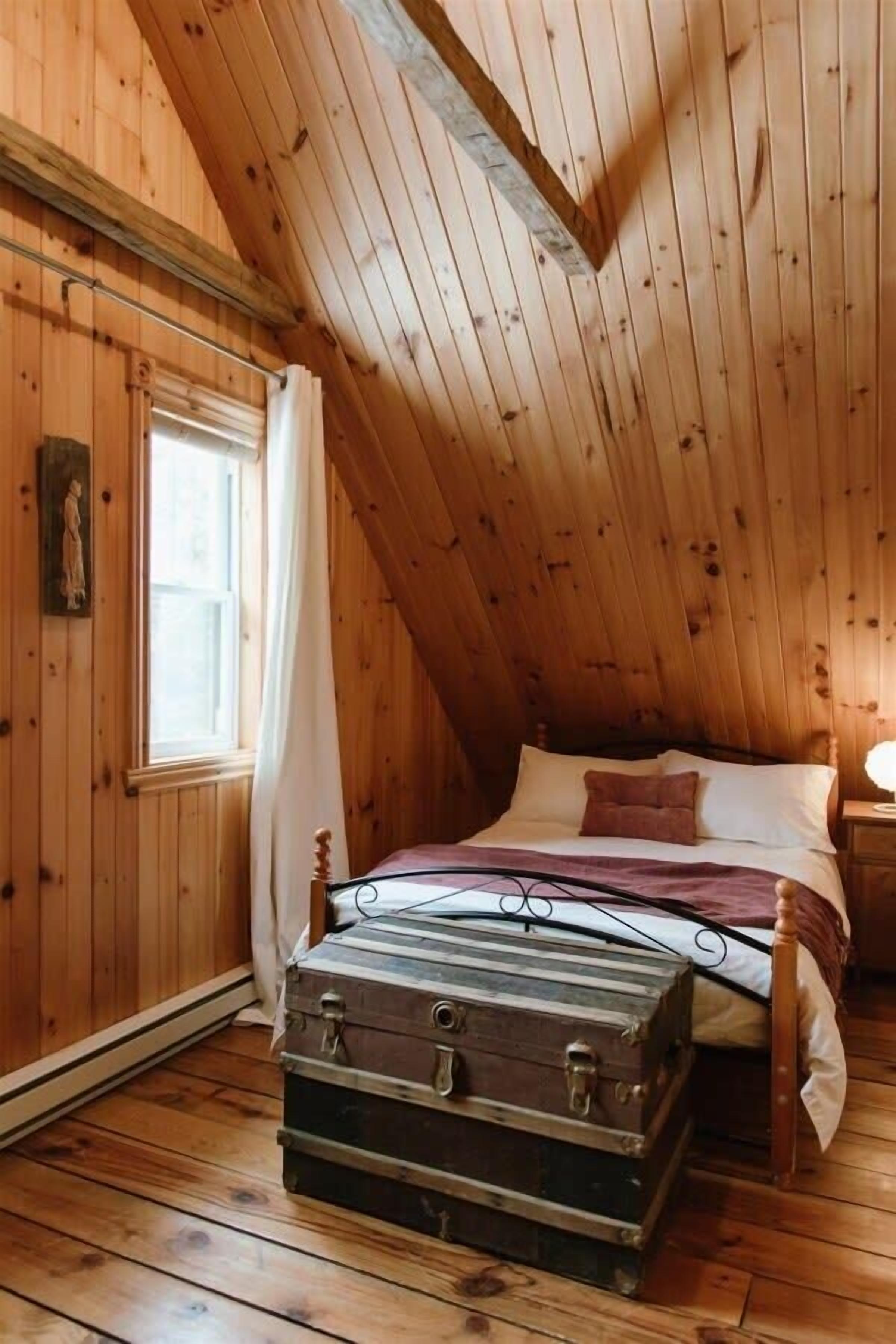 A wooden-paneled room with a sloped ceiling, a bed with a metal frame, a wooden chest, and a window with curtains.