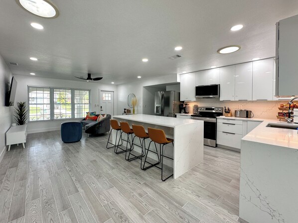 A modern kitchen with a central island, bar stools, a refrigerator, microwave, oven, and a large window.