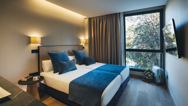 Una habitación de hotel moderna con una cama grande, una mesita de noche con una lámpara, una pequeña mesa con un libro y una taza, y una ventana con cortinas.
