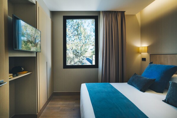 A hotel room with a bed, a television mounted on the wall, a window with curtains, and a bedside lamp.