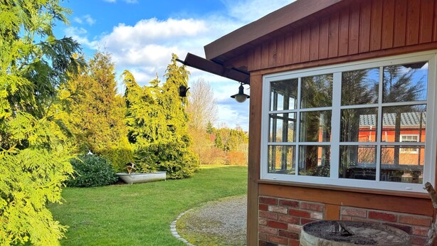Een houten gebouw met een groot raam, een kleine tuin met gras en bomen, en een houten vat.