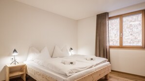A minimalist bedroom with a wooden bed, two bedside tables with lamps, a window with blinds, and a plain white wall. - HUUS123, Apartment 3 (Au)