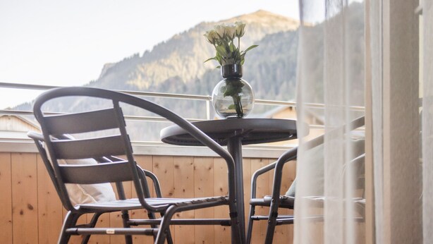 Un balcon avec une table et des chaises, un vase rempli de fleurs et un paysage montagneux en arrière-plan.