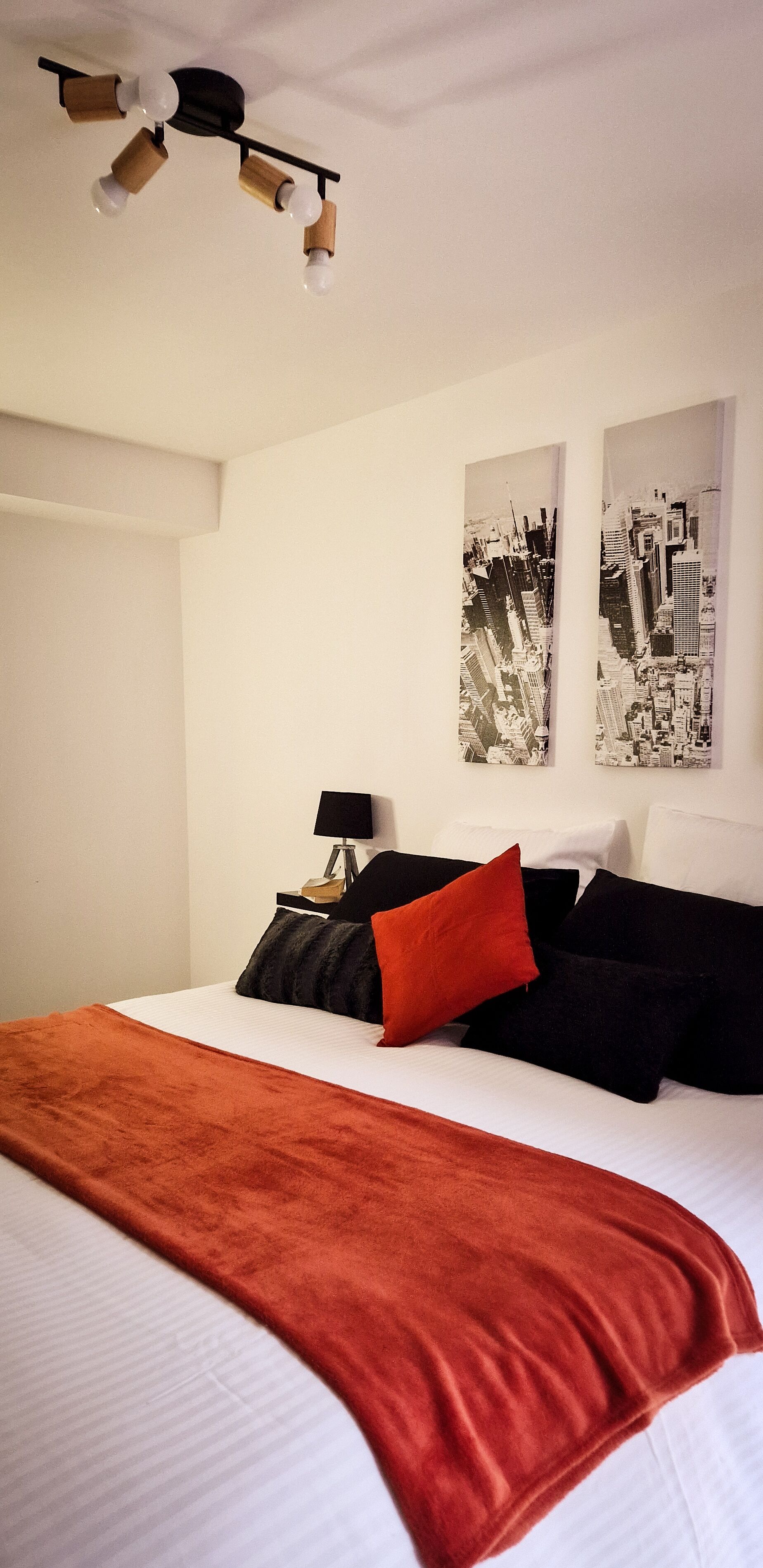 A hotel room with a bed, a red throw blanket, black and red pillows, a nightstand with a lamp, and two black-and-white cityscape paintings on the wall.