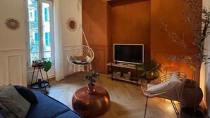 Olohuoneessa on sohva, jalustalla oleva televisio, riipputuoli ja ruukussa oleva kasvi.