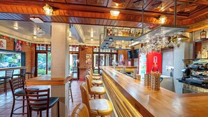 Un espace bar avec des tabourets de bar en bois, un comptoir avec une caisse enregistreuse, et une variété de bouteilles et de verres exposés sur des étagères.