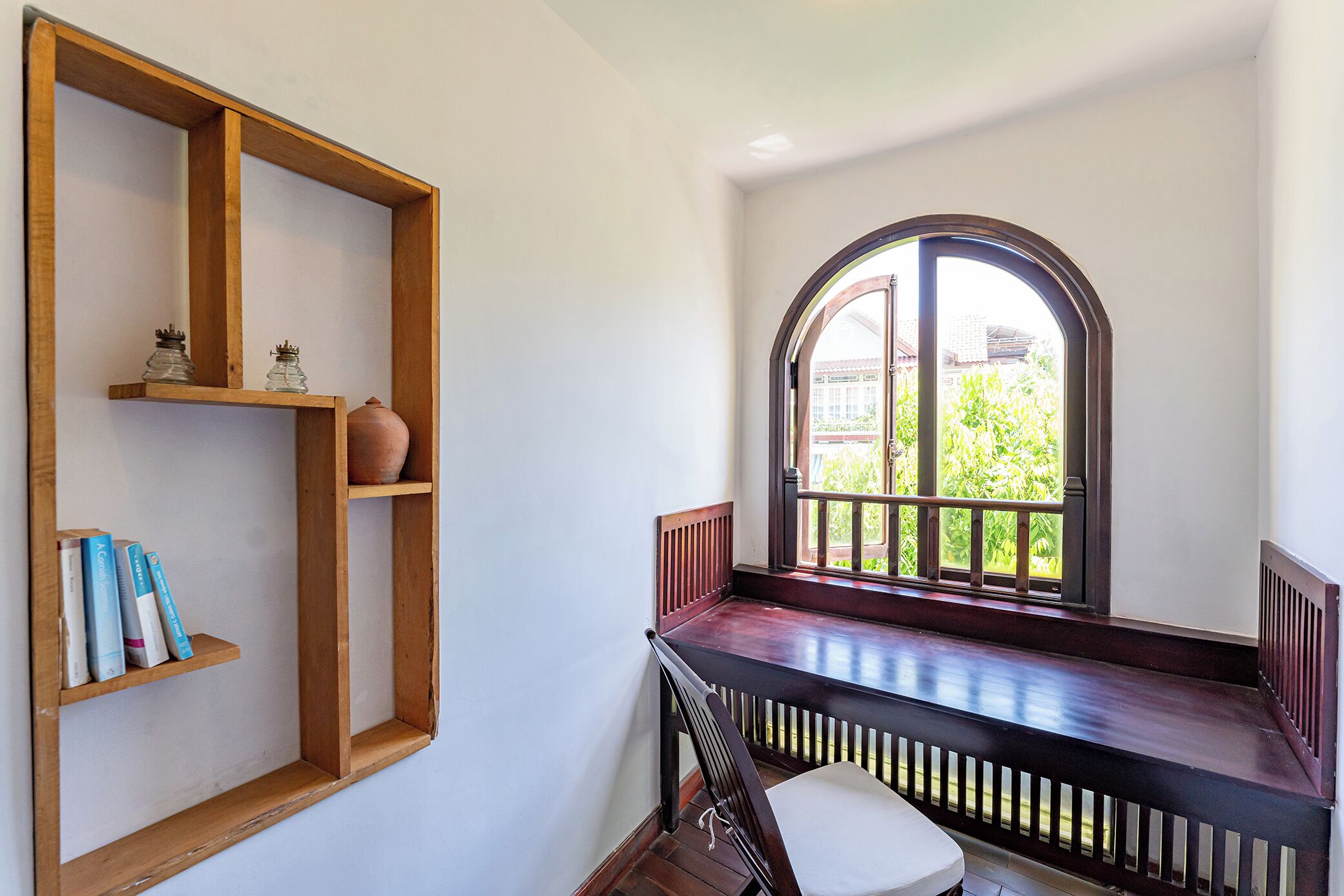 A room with a wooden bench, a chair, a bookshelf, and a large arched window with a view of greenery outside.