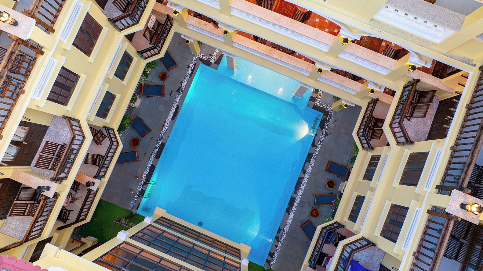 An aerial view of a hotel with a central swimming pool surrounded by balconies and outdoor seating areas.