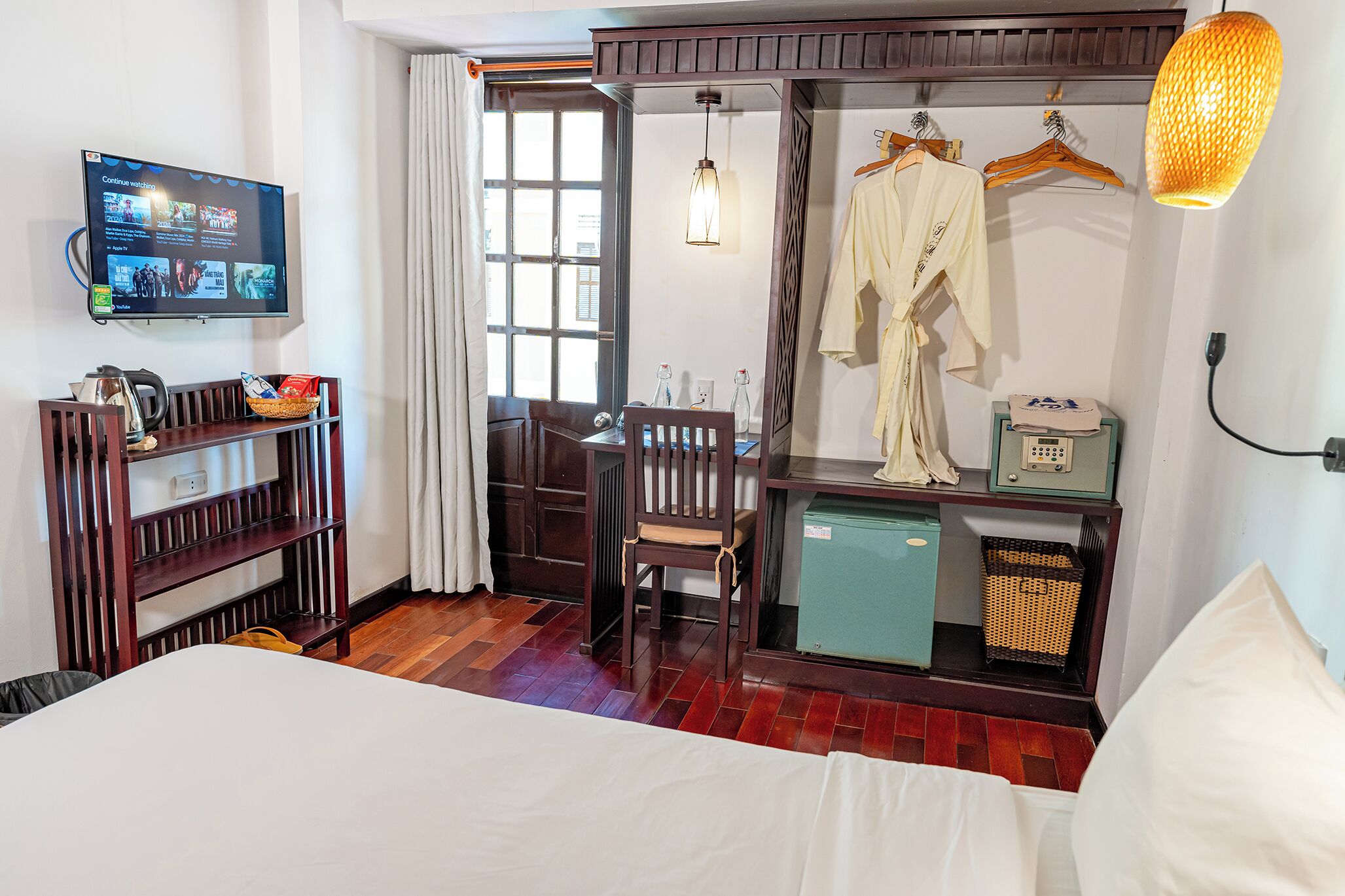 Una habitación de hotel con cama, escritorio y silla, televisor montado en la pared, ventana y armario con ropa colgada.