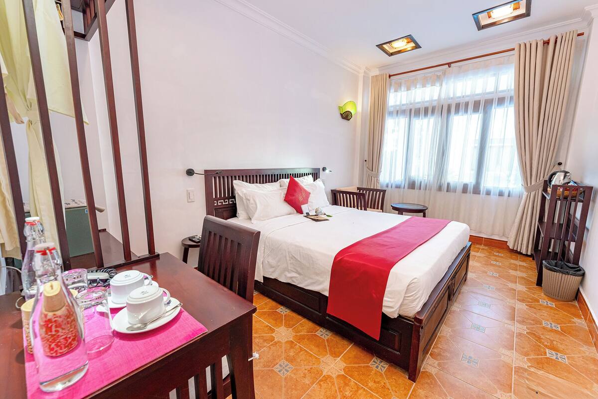 Ein Hotelzimmer mit einem Bett, einem kleinen Tisch mit Teeservice, einem Sessel, einem Fenster mit Vorhängen und einer Wandleuchte.