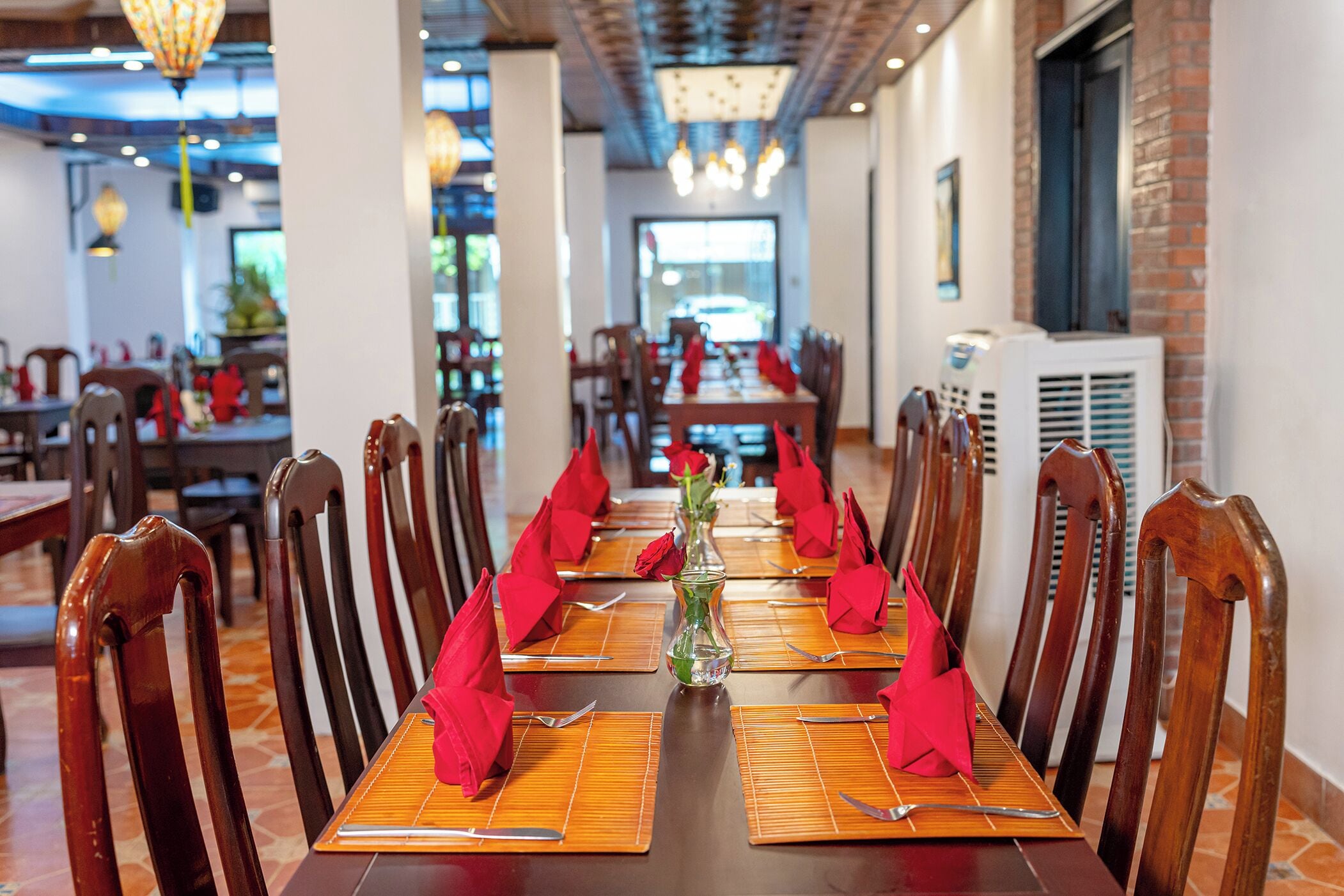 Una larga mesa de comedor dispuesta con servilletas rojas, cubiertos y un florero con flores.