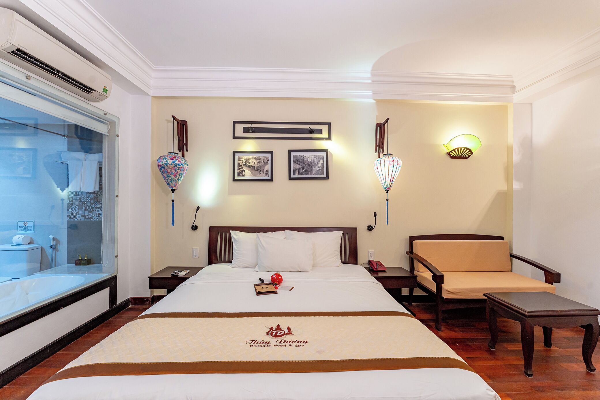 Quarto de hotel com uma cama grande, mesa de cabeceira com abajur, uma cadeira, uma mesinha e prateleira na parede com objetos decorativos.