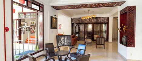 A spacious lobby with a high ceiling, a ceiling fan, and a traditional wooden partition. There are several seating areas with chairs and tables, and a large window offering a view outside.