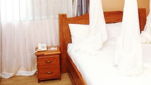 Un dormitorio con cama de madera, ropa de cama blanca, una mesita de noche con un teléfono y una ventana con cortinas traslúcidas.
