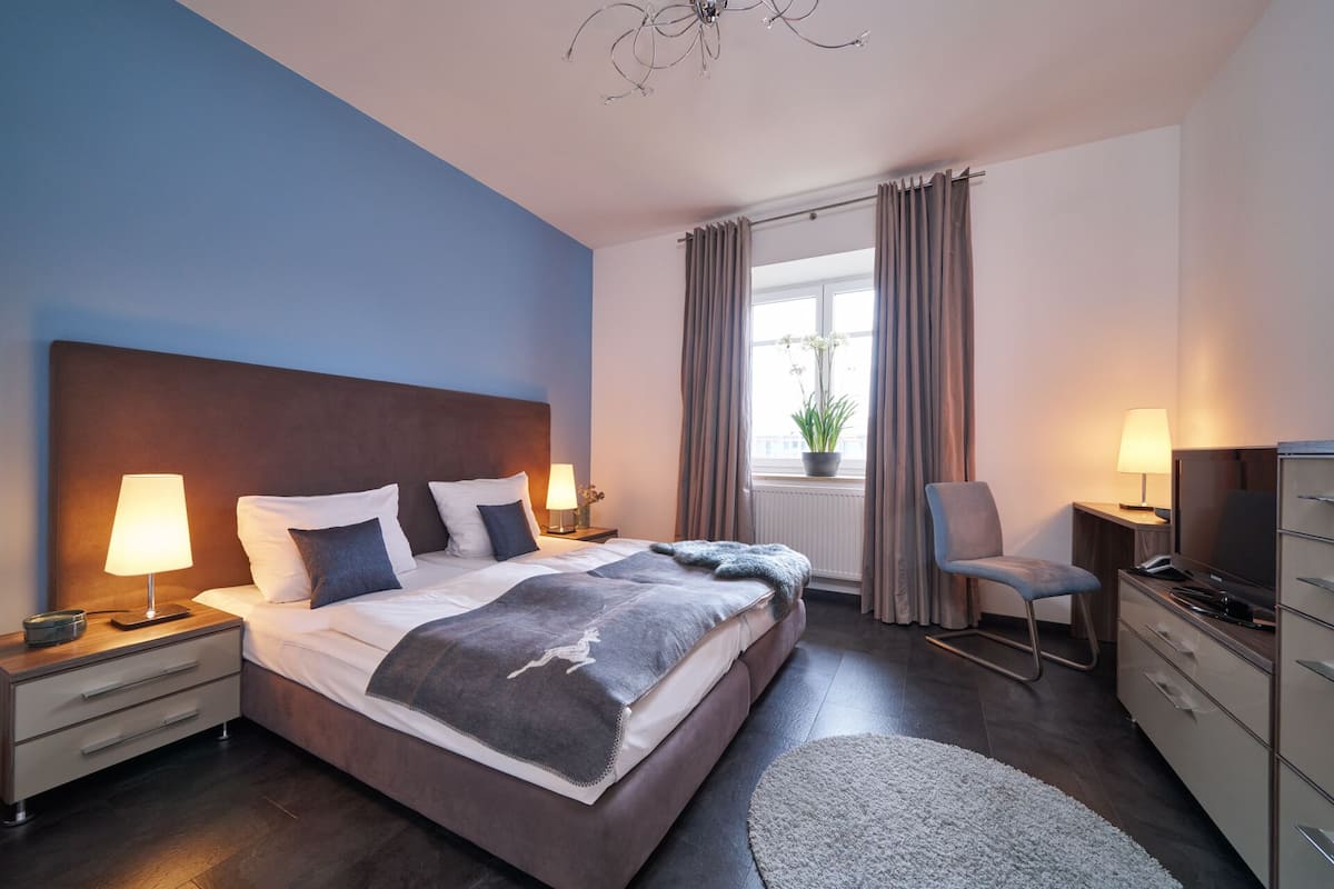 A modern hotel room with a large bed, bedside lamps, a desk with a chair, a TV, and a window with curtains.