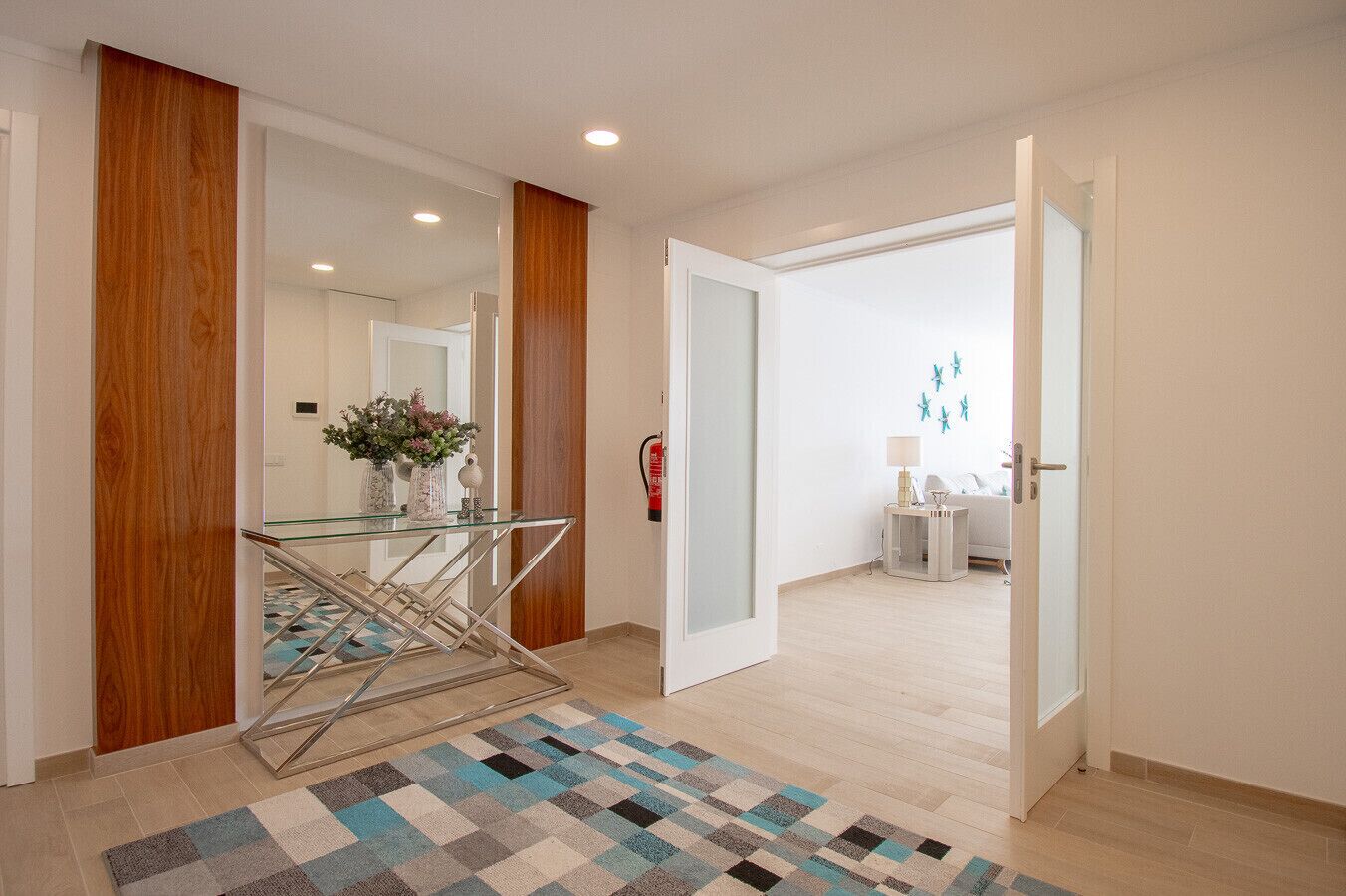 A modern hotel corridor with a glass table, a patterned rug, wooden accents, and a visible bathroom with a sink and mirror.