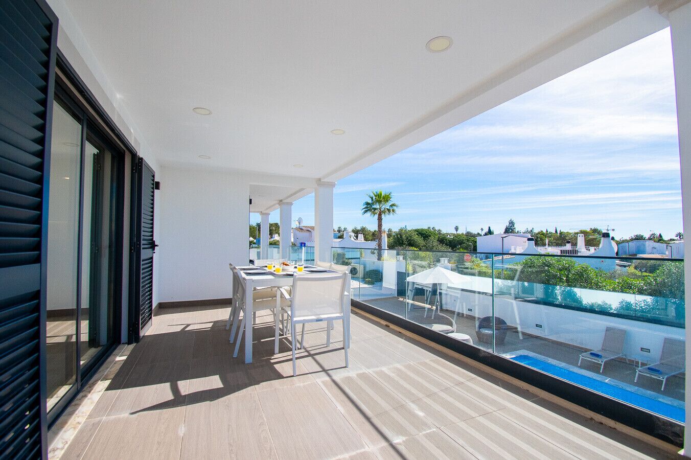 A balcony with a table and chairs, a glass railing, and a view of buildings and trees.