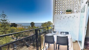 A balcony with a table set for dining, overlooking a scenic view of the ocean and a parking area.