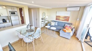 A modern apartment with a kitchen, dining area, and living room. It includes a glass dining table, a sofa, and a TV.
