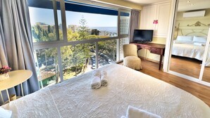 A hotel room with a large bed, a desk with a TV, and a view of the city and sea.