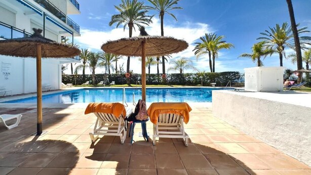 A poolside area with lounge chairs and umbrellas, a pool, palm trees, and a building in the background.