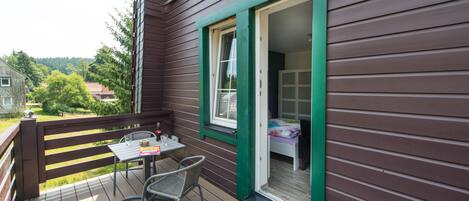 Une terrasse en bois avec une table et des chaises, une fenêtre à cadre vert et une vue sur une zone boisée.