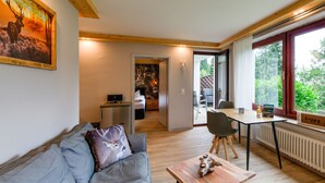 En moderne stue med sofa, sofabord og et spiseområde med bord og stole. Et maleri hænger på væggen, og et vindue viser udsigt til grønne omgivelser.