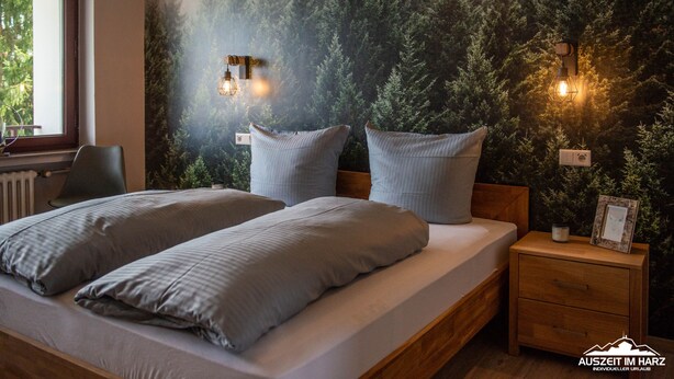 Une chambre à coucher avec un lit, deux oreillers, une table de chevet, une chaise et un mur orné d’une fresque représentant une forêt.