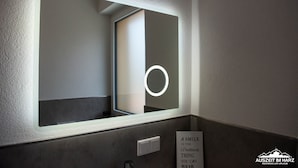 Une salle de bain avec un grand miroir, un luminaire au-dessus et un panneau affichant une citation.