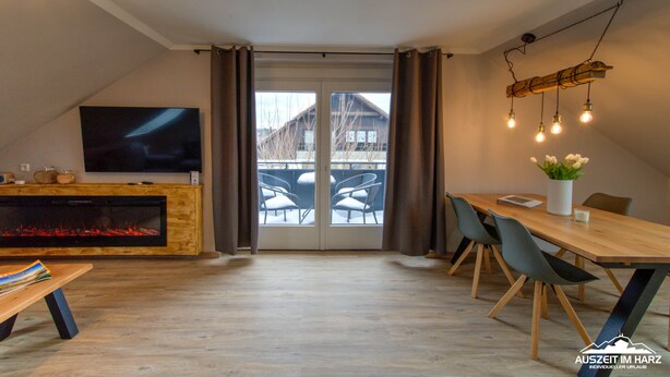 A modern living room with a fireplace, a dining table with chairs, a TV, and a balcony with chairs.