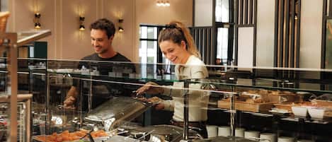 Zona de bufé con diferentes alimentos en recipientes transparentes, un empleado sirviendo comida y un interior moderno con iluminación colgante.