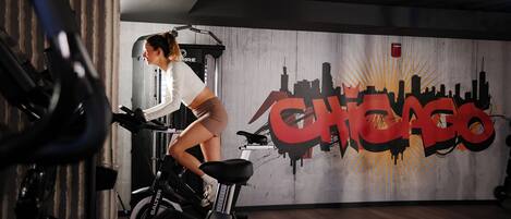 A woman cycling on a stationary bike in a gym with a mural of the Chicago skyline in the background.