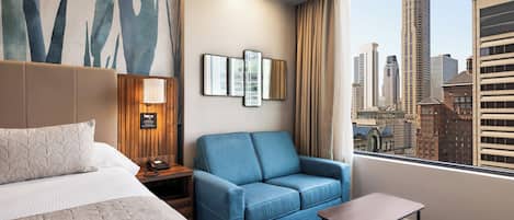 A hotel room with a bed, a blue sofa, a small table, a nightstand, and a city view.