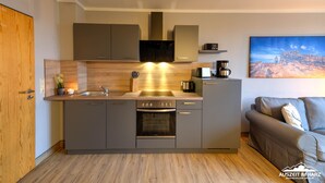 Una cocina moderna con horno empotrado, cafetera y lavaplatos. A la izquierda hay una puerta de madera y a la derecha un sofá gris.