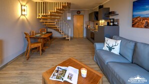 Una sala de estar moderna con una escalera de madera, una zona de comedor, una cocina y un sofá gris con un cojín con forma de ciervo.