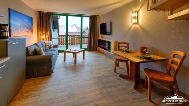 Una sala de estar moderna con sofá, mesa de centro, mesa de comedor y sillas. Hay un televisor montado en la pared y una chimenea. La habitación tiene ventanas grandes y un balcón.