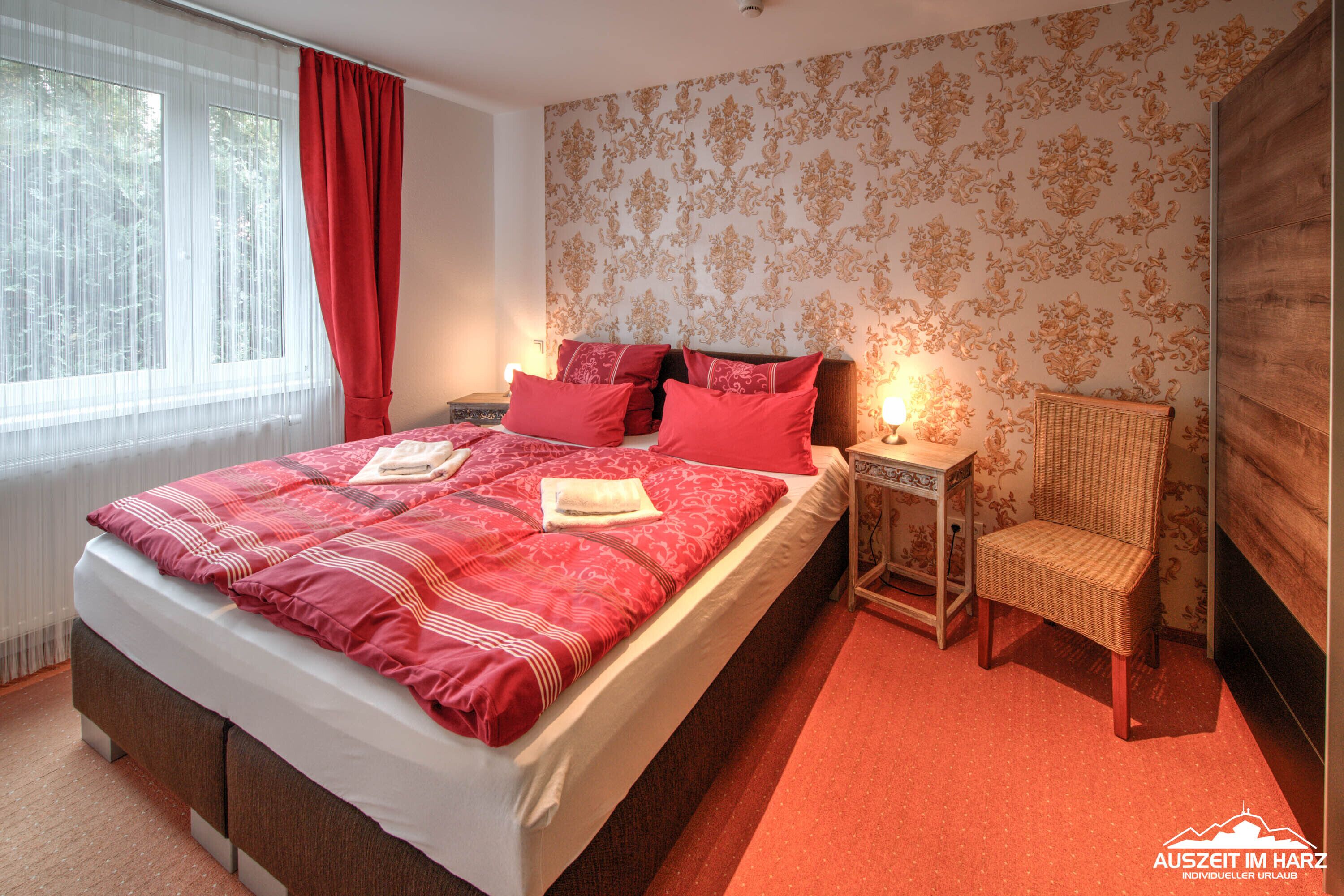 A bedroom with a large bed, red bedding, a nightstand with a lamp, a wicker chair, and a radiator.