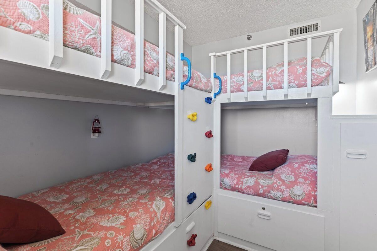 Une chambre avec un lit superposé, composé d’un lit du bas et d’un lit du haut, tous deux dotés de draps à motifs floraux. Des crochets colorés sont fixés au mur pour ranger les affaires.