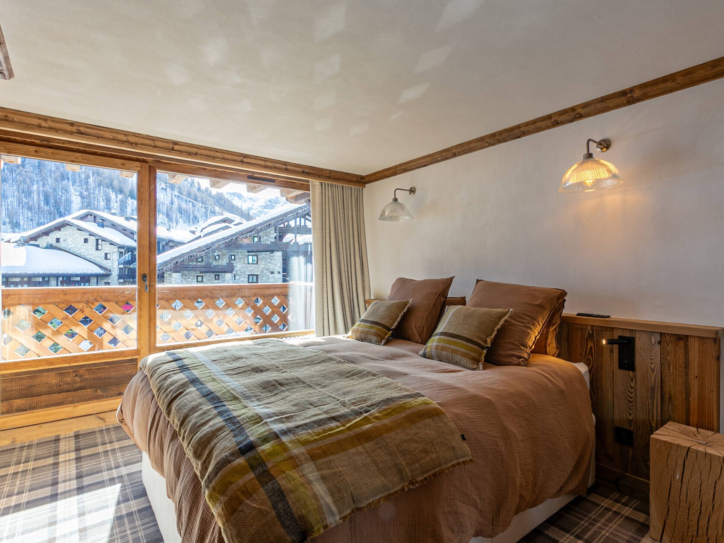 A bedroom with a large bed, wooden headboard, and a view of snow-covered buildings through a balcony.