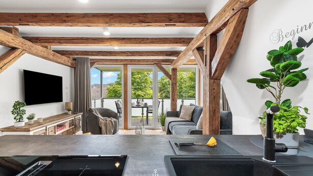 Eine moderne Küche mit schwarzem Arbeitsplatz und Spüle, Holzbalken an der Decke und einem großen Fenster mit Blick auf Grünflächen.